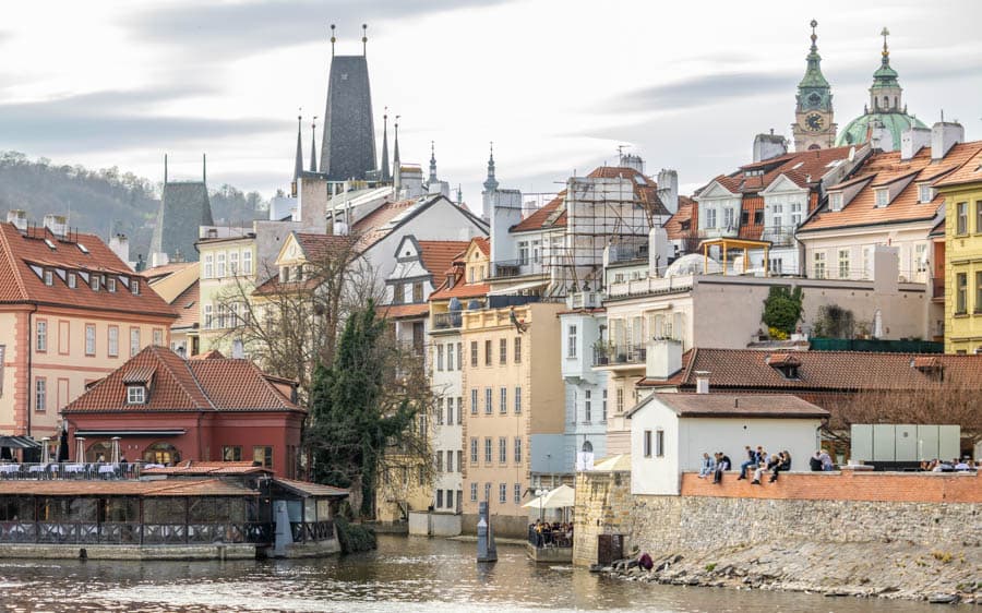 伏爾塔瓦河岸景致，重點展示了小城區（Malá Strana）與查理大橋附近的景色，照片右側牆面上方為小城區碼頭的一部分，遠處可見馬拉斯特蘭斯克廣場（Malostranské náměstí）附近的建築天際線。